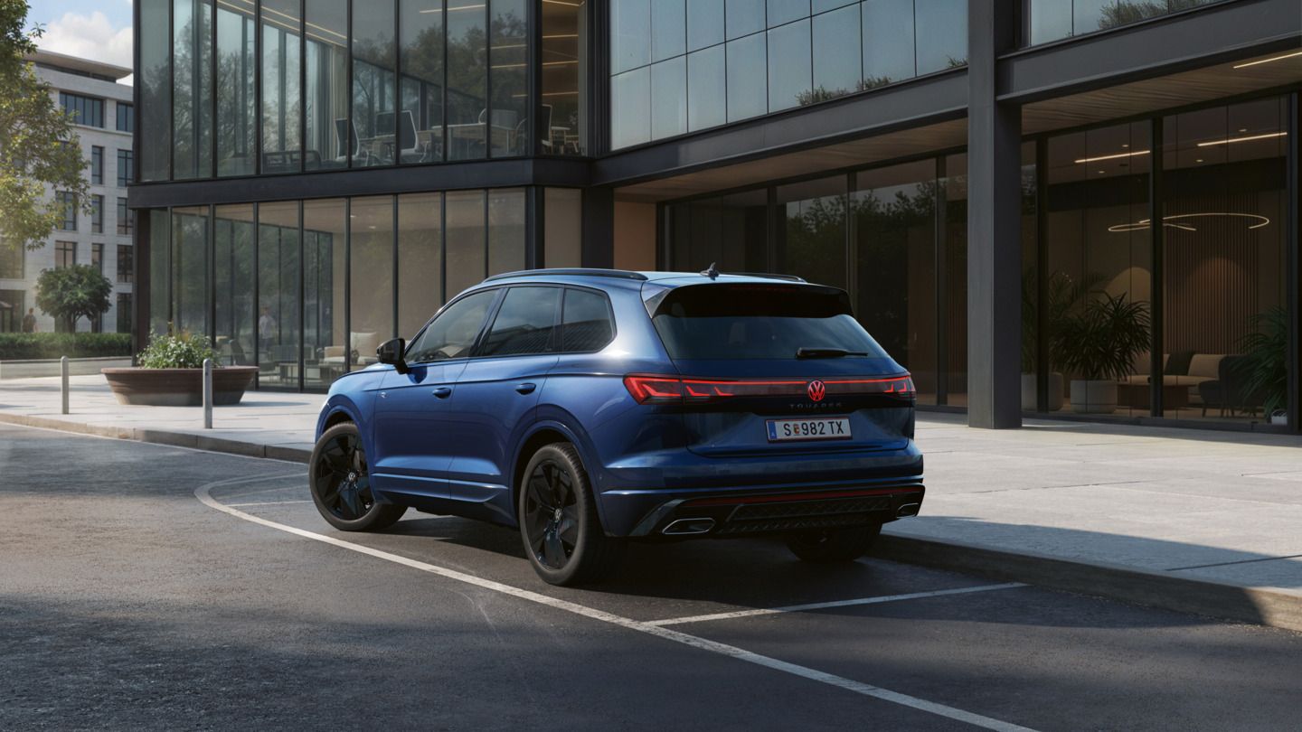Ein blauer VW Touareg FINAL EDITION  steht vor einem Bürogebäude mit Glasfassade - Ansicht von schräg hinten.