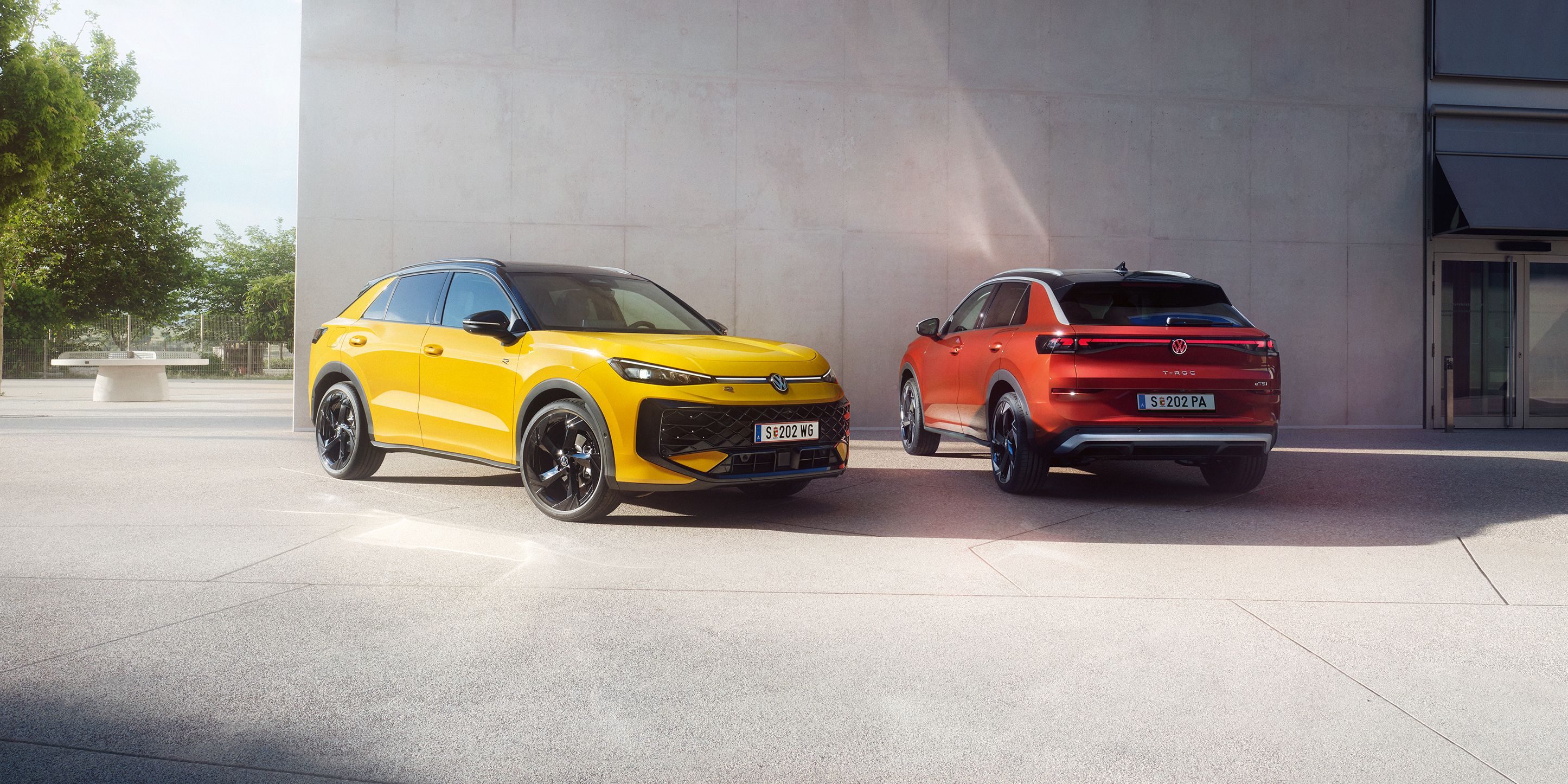 Ein gelber VW T-Roc R-Line und ein roter VW T-Roc Style stehen nebeneinander vor einem modernen Gebäude.