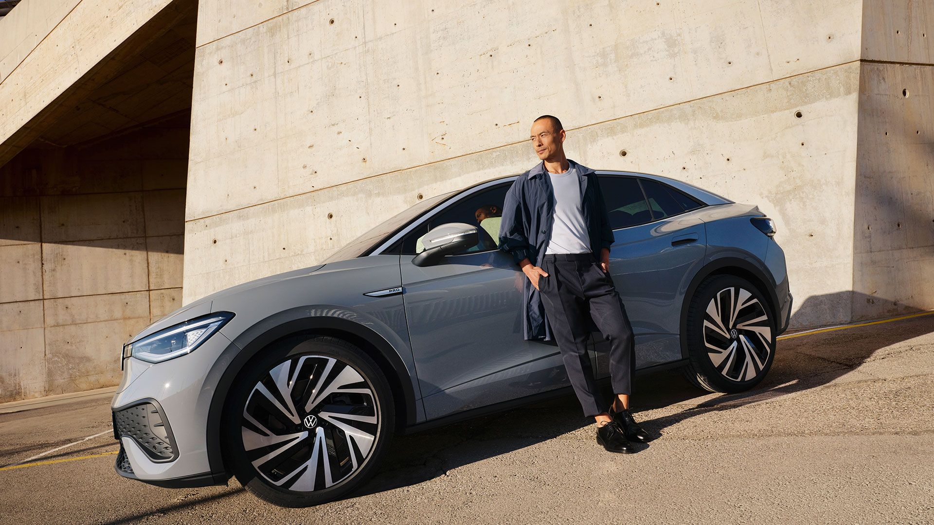 Seitenansicht eines grauen VW ID.5 vor einer hellen Wand, ein Mann lehnt sich an die Fahrertür an.