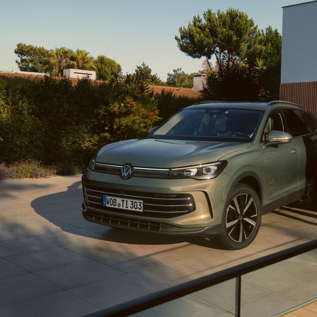 Grüner VW Tiguan parkt in Frontansicht auf der Einfahrt eines modernen Wohnhauses. Ein junges Paar steht daneben und umarmt sich.