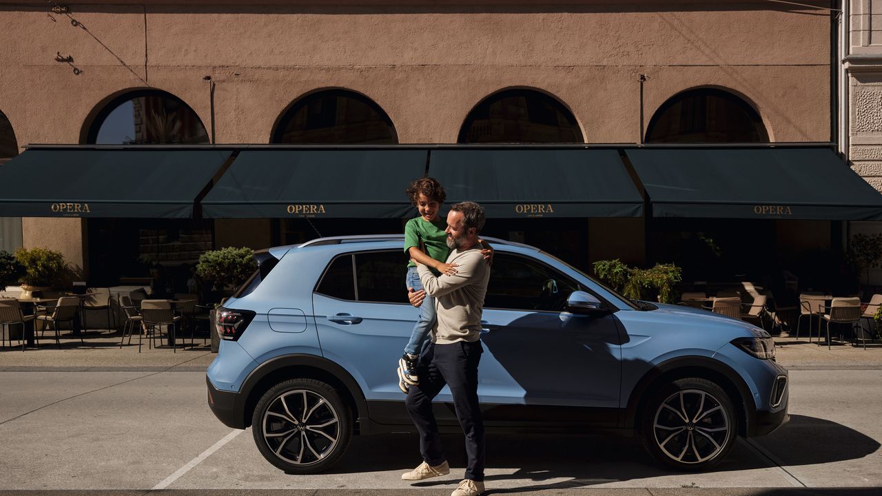 Ein Mann hält einen fröhlichen Jungen vor einem blauen VW T-Cross auf dem Arm.