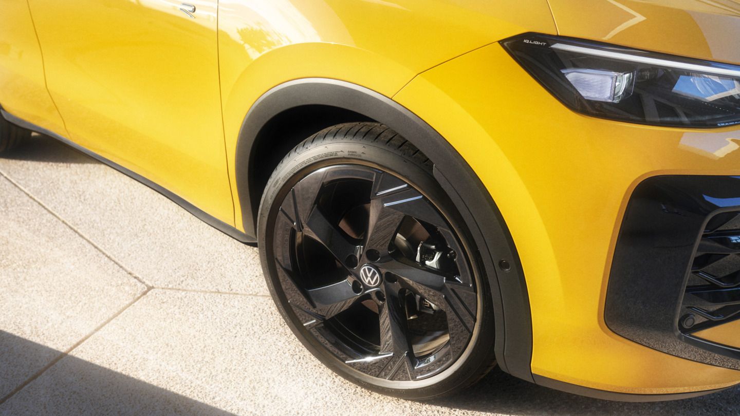 Das Bild zeigt einen Teil eines gelben T-Roc aus der Seitenansicht. Der Fokus liegt auf dem schwarzen Vorderrad und der Kotflügel. Das Auto steht auf hellen, rauen Bodenplatten.