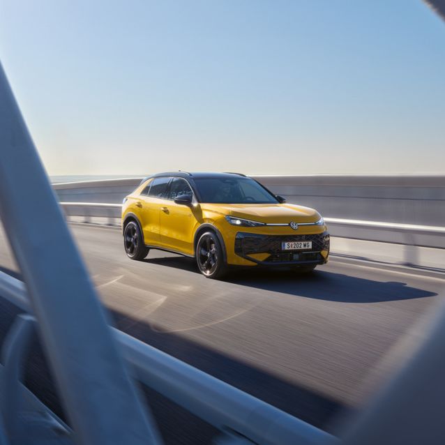 Ein gelber T-Roc fährt über eine moderne Brücke bei klarem Himmel. Die Aufnahme zeigt das Fahrzeug aus der Ferne, teilweise durch die Brückenstrukturen hindurch. Es ist ein sonniger Tag.