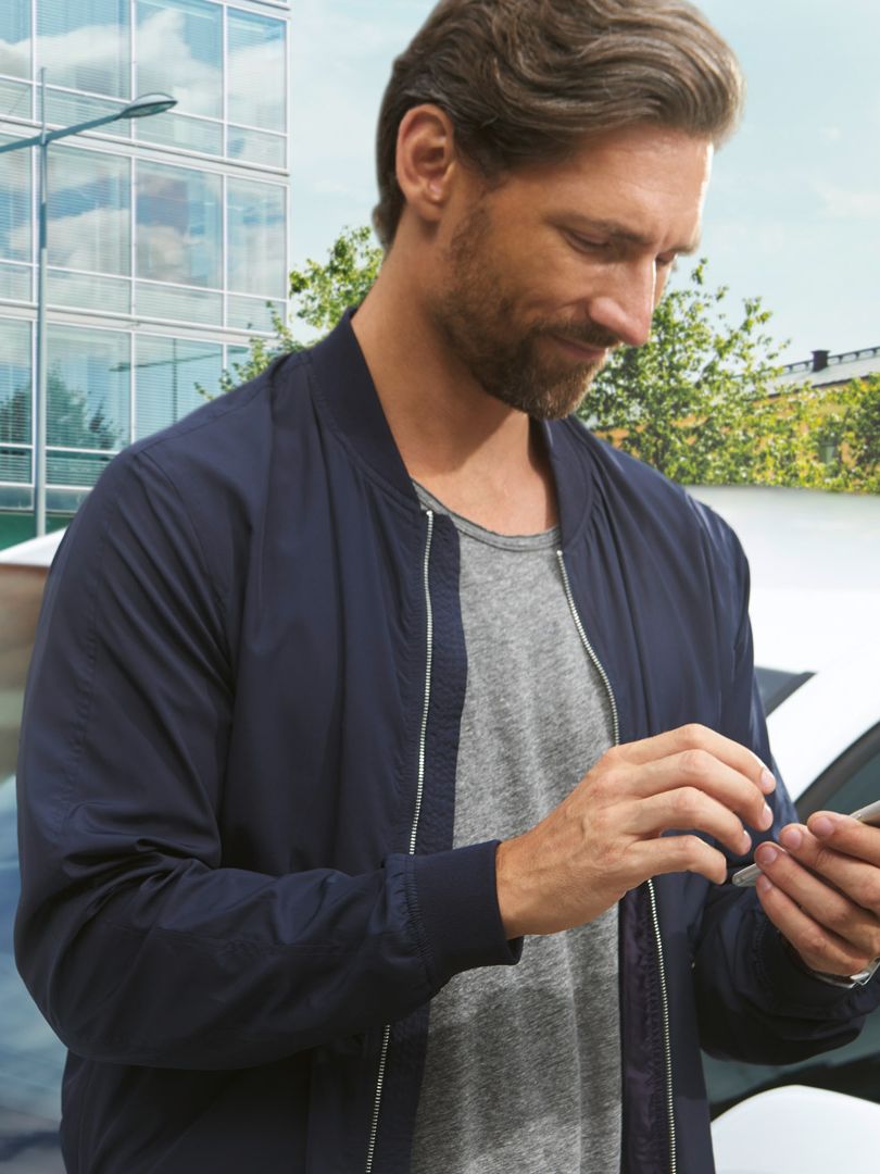 Ein Mann tippt in sein Smartphone. Im Hintergrund ein weißer Volkswagen vor einem Gebäude.